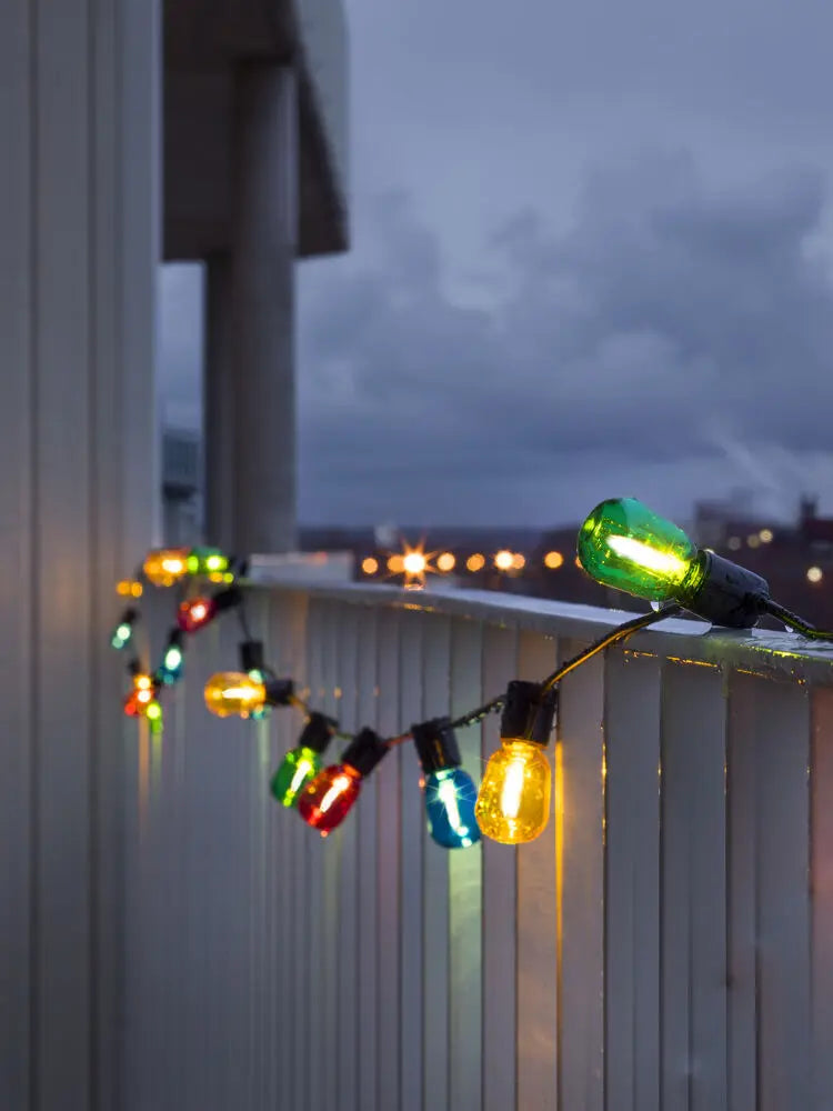 Konstsmide lichtsnoer gespannen over een houten frame boven een loungebed buiten, verlicht in de avondschemering