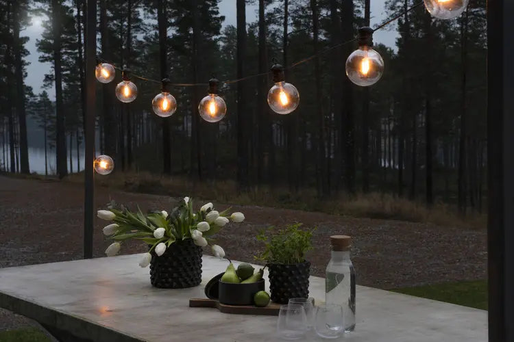 Konstsmide lichtsnoer opgespannen boven een terras met tafel, verlicht bij avondlicht met bos op de achtergrond