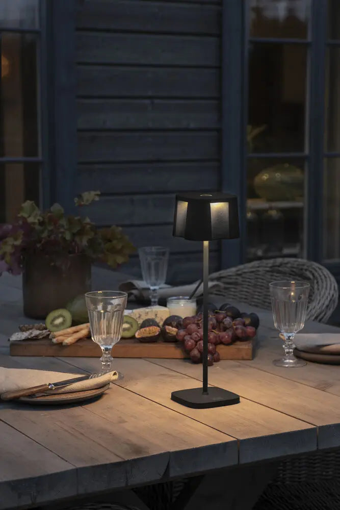 Konstsmide Nice tafellamp op een gedekte tafel, verlicht bij avondlicht met een blauwe gevel op de achtergrond