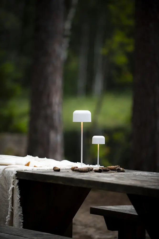 Witte Konstsmide Lille tafellamp op een houten tafel, verlicht tussen bomen