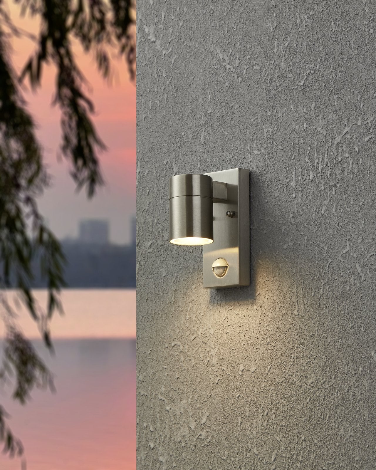EGLO Riga 5 wandlamp gemonteerd aan een gevel, verlicht bij avondlicht met bladeren op de voorgrond